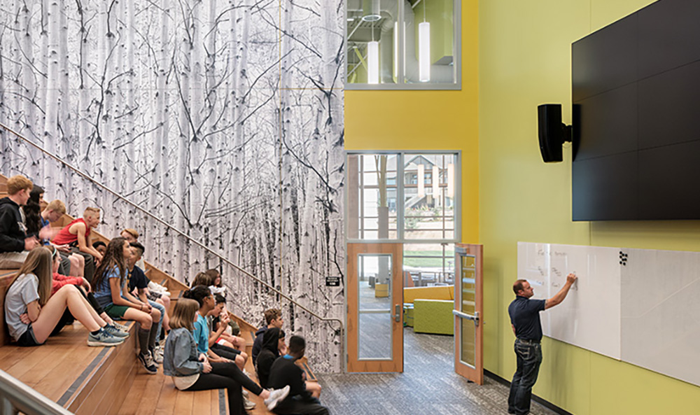 A teacher writes on a whiteboard in front of a group of middle school students seated on a learning stair