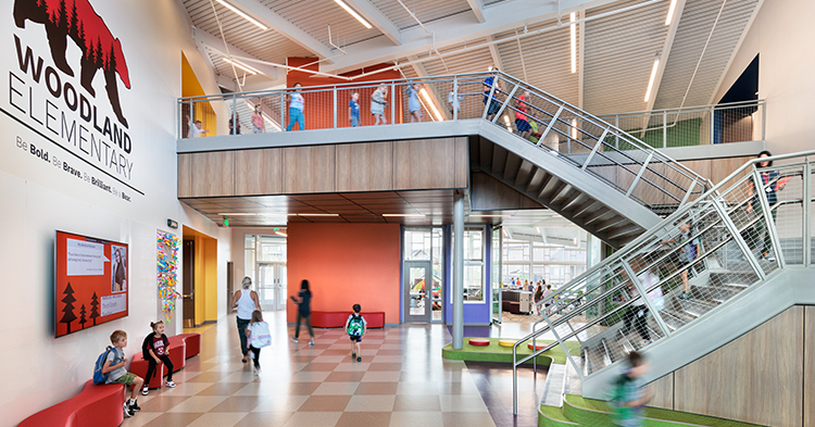 a colorful hallway at woodland elementary school with a staircase leading to a second story; several young students are sitting on benches and walking in the hallway.